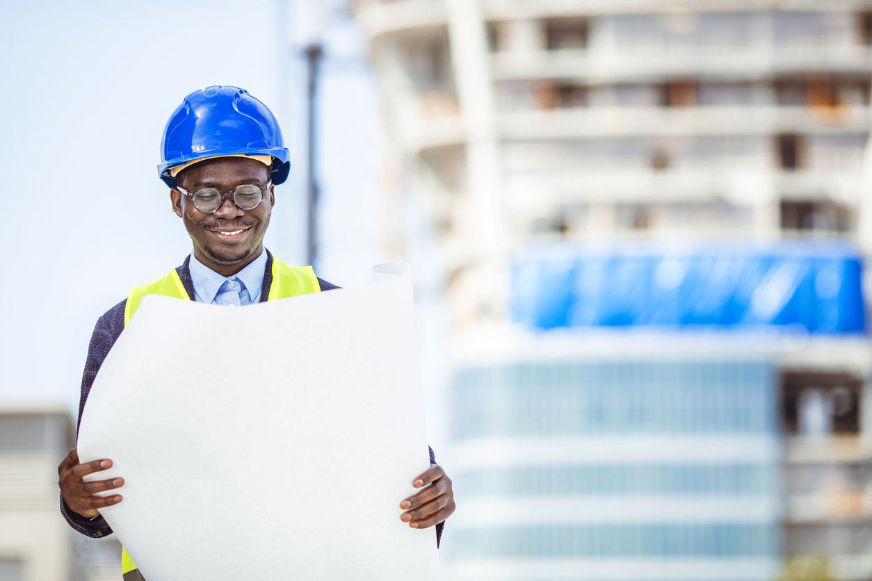 Engineer on site checking project. Portrait of smiling young architect or handsome engineer holding rolled up blueprints with building construction background.