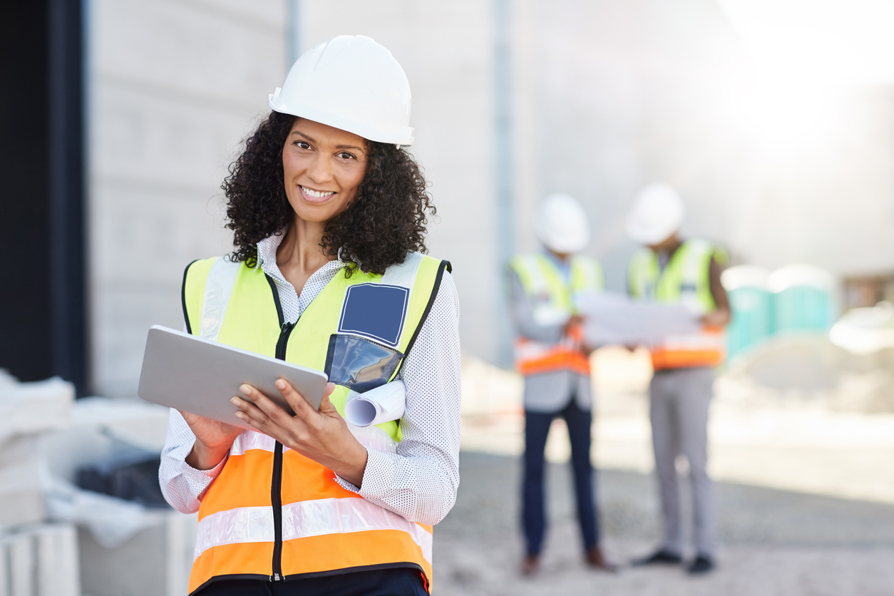 Portrait of a smiling female construction engineer wearing a safety vest standing on a worksite using a digital tablet with workers in the background