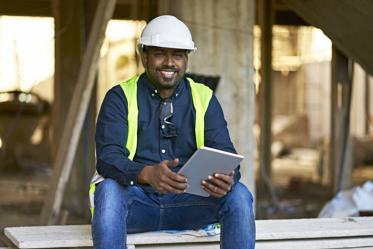 Portrait of male engineer holding digital tablet at construction site. Smiling architect is sitting on planks. He is wearing protective workwear.